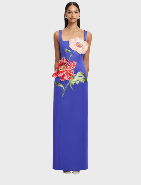 A woman in a blue dress stands gracefully, showcasing her stylish outfit. The dress features elegant shoulder details and falls stylishly around her figure.