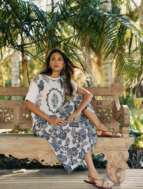 A woman in a white shirt and blue floral skirt sits on a wooden bench, enjoying her surroundings.