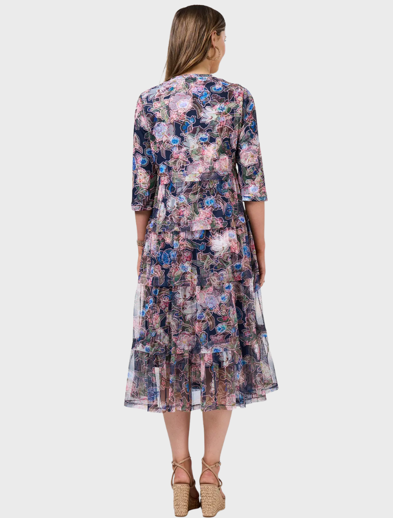A woman in a floral dress stands smiling, showcasing the vibrant patterns of her attire. The dress features colorful flowers against a light background.
