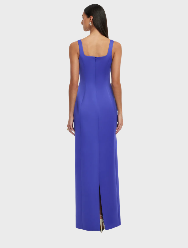 A woman wearing a blue dress, standing confidently. The dress features a stylish design and fits her shoulders elegantly. She appears poised, showcasing the fabric's texture and pattern in a bright daylight setting.