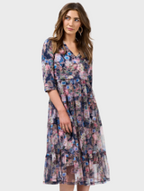 A woman wearing a dress with a unique pattern, showcasing her long hair. She is standing casually and has a friendly facial expression.