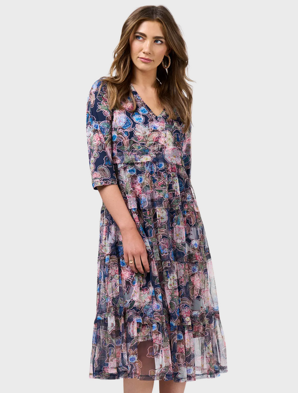 A woman wearing a dress with a unique pattern, showcasing her long hair. She is standing casually and has a friendly facial expression.