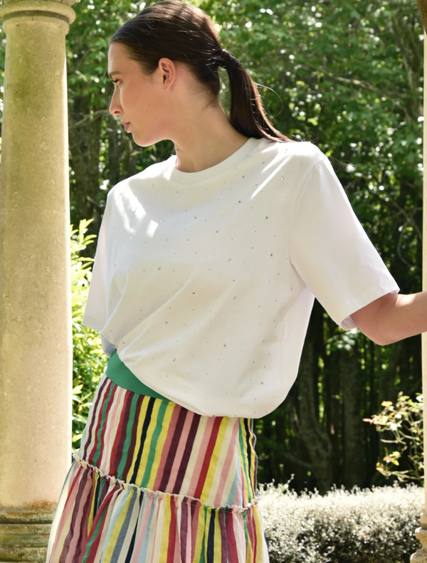 A woman wearing a white Sparkle Magic T-shirt and a multicolored striped skirt stands confidently, smiling at the camera.