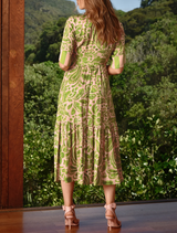 A woman dressed in green and wearing heels stands by a window, framed by soft light filtering through the glass.