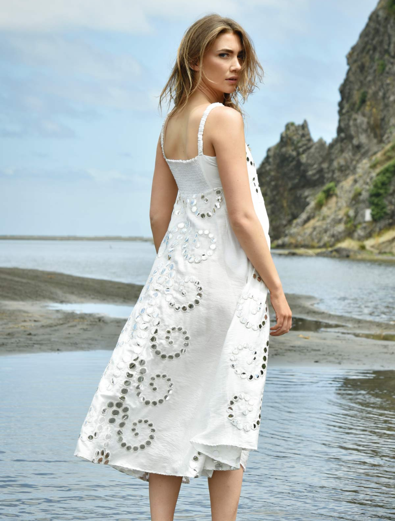A woman in a white dress stands on the shore, with the ocean waves and a bright sky in the background.