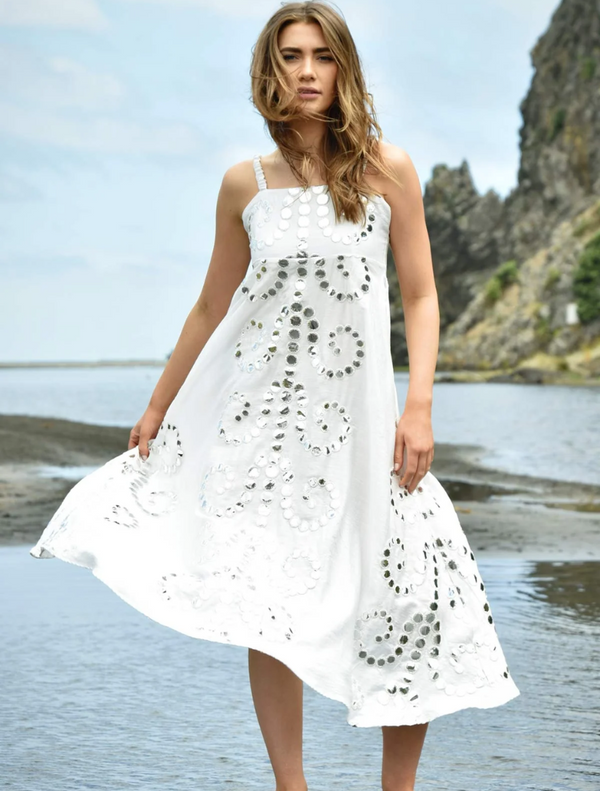 A woman in a flowing white dress stands gracefully on the beach, with waves gently lapping at her feet.
