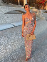 A woman in a colorful dress stands on a patio, holding an Amalia Pleated Woven Clutch in natural tones.