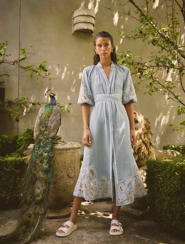 A woman in a blue embroidered maxi dress stands beside a vibrant peacock, showcasing elegance and nature's beauty.