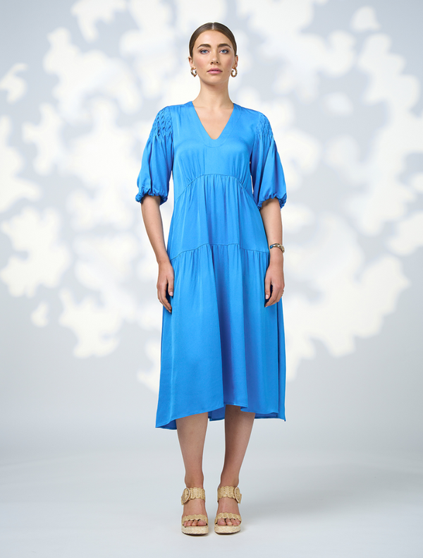 A model is dressed in a blue v-neck dress from Genf Gilet - Candy, presenting a sophisticated and fashionable look.