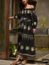 A woman dressed in black leans casually against a wall, showcasing a stylish and relaxed demeanor.