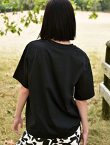 A woman wearing a black Sparkle Magic T-shirt and black pants stands in a grassy field, looking confidently ahead.