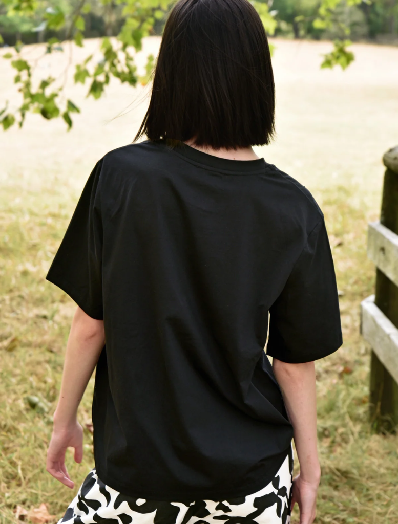 A woman wearing a black Sparkle Magic T-shirt and black pants stands in a grassy field, looking confidently ahead.