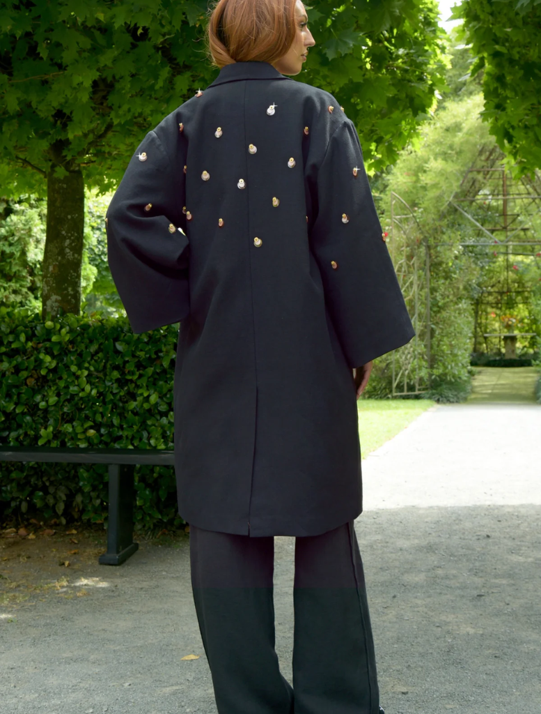 A woman in a black coat and pants stands confidently in a park, surrounded by greenery and trees.
