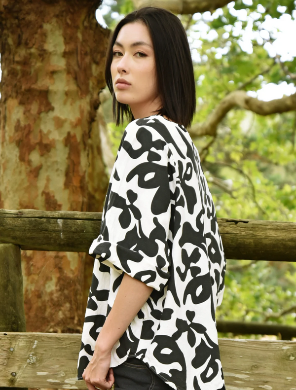 A woman wearing a black and white patterned top stands casually against a fence, with a relaxed expression.
