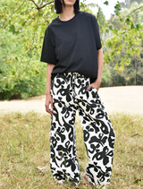 A woman dressed in black and white pants leans against a fence, exuding a laid-back vibe in an outdoor setting.