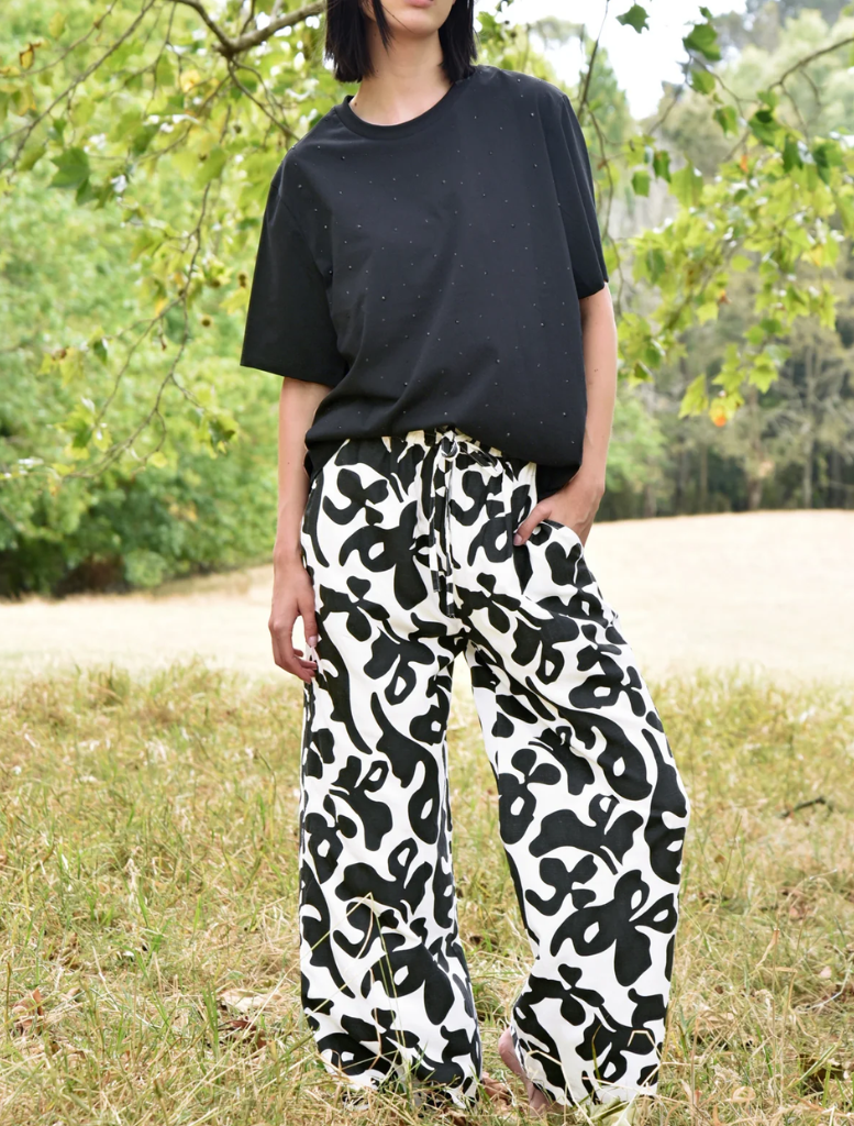A woman dressed in black and white pants leans against a fence, exuding a laid-back vibe in an outdoor setting.