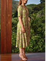 A woman wearing a green dress and heels poses by a window, showcasing her stylish outfit in soft, natural light.