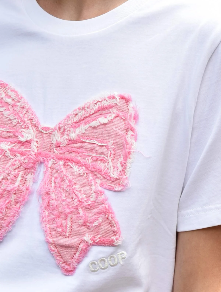 A woman showcases a pink skirt paired with a white t-shirt that has a bow, exuding a cheerful demeanor.