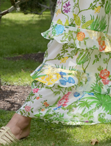 A woman in a vibrant floral dress walks through a park, with trees and flowers in the background.
