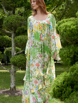 A woman in a vibrant floral dress walks through a park, with trees and flowers in the background.