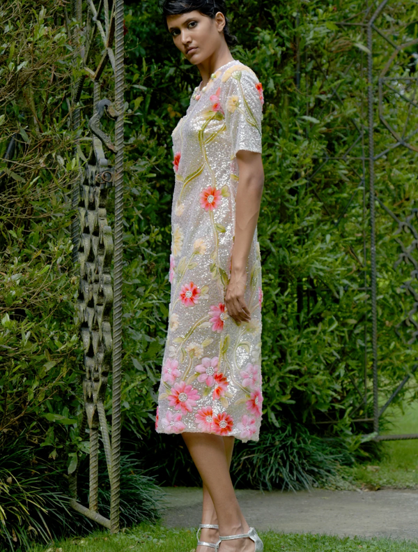 A woman wearing a silver and pink floral dress rests against a tree, surrounded by a peaceful outdoor setting.