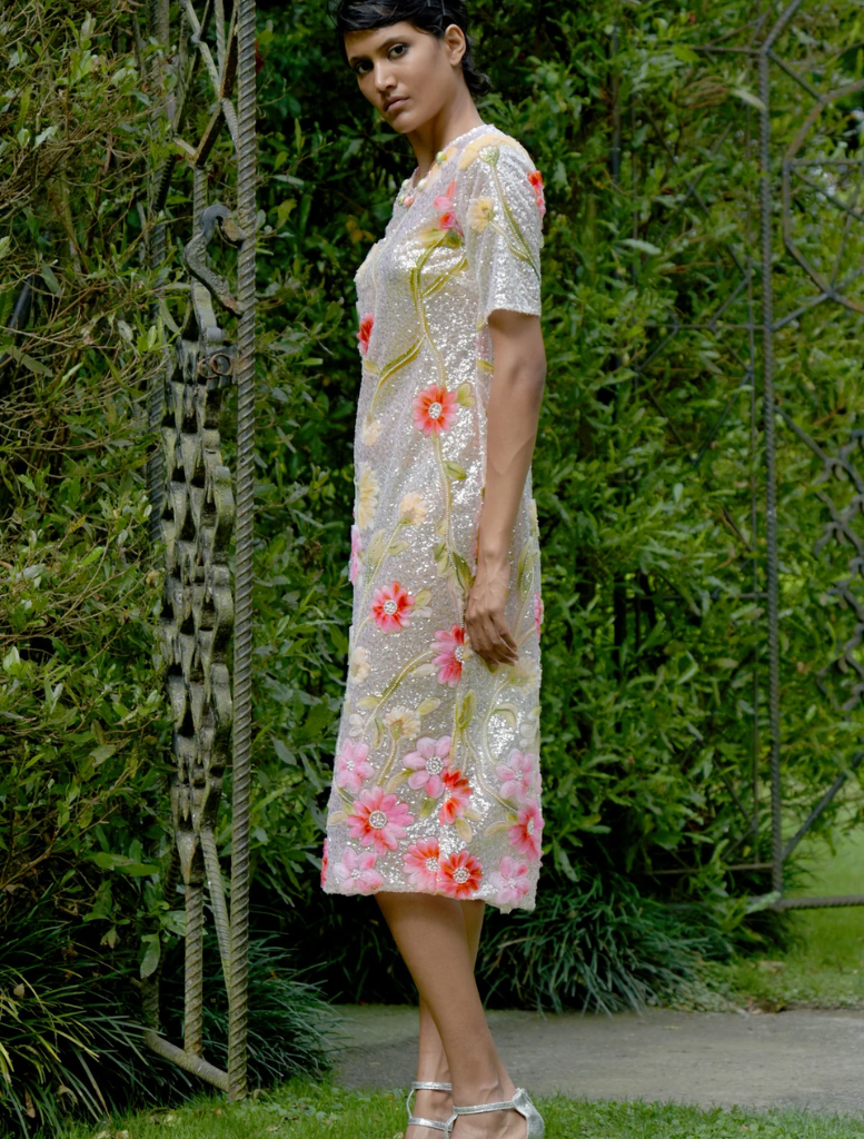 A woman wearing a silver and pink floral dress rests against a tree, surrounded by a peaceful outdoor setting.