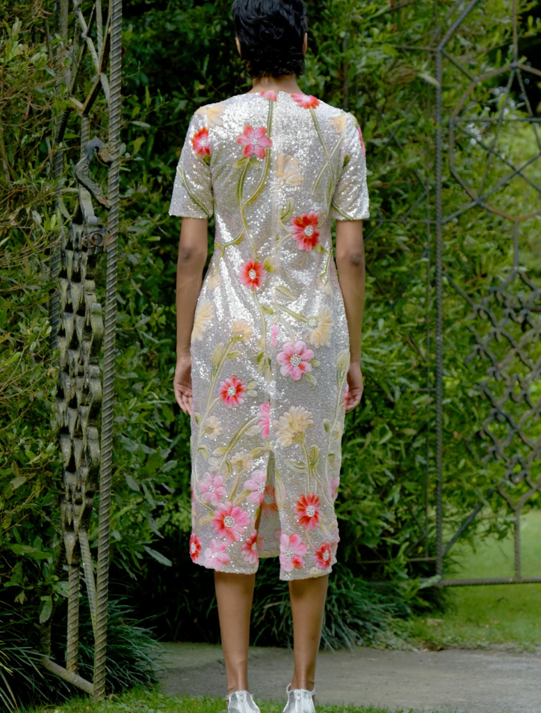 A woman in a silver and pink floral dress stands gracefully in a vibrant garden filled with blooming flowers.