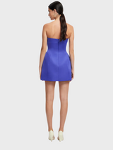 Back view of a woman wearing a flowing blue dress, standing gracefully against a neutral background.