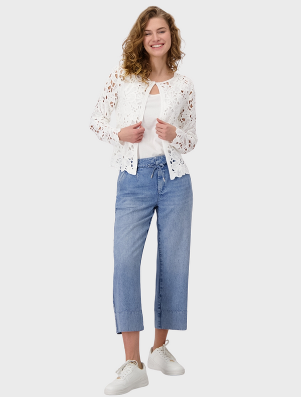 A woman in a white lace top and blue jeans stands confidently, showcasing her stylish outfit.