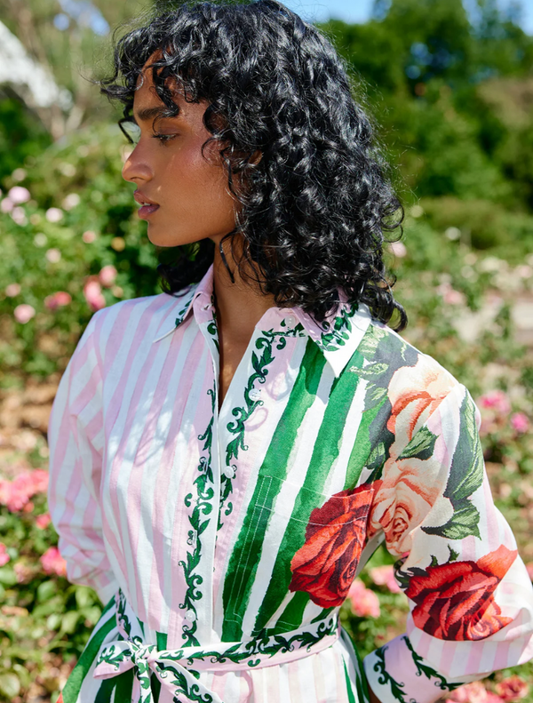 A woman wearing a floral dress, the Demi Dress in Leaf Rose Multi, poses in a lush garden filled with colorful flowers.