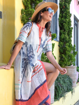 A woman wearing a colorful dress and hat sits on a step, showcasing the vibrant 'Don't Make Waves Dress - Marina.