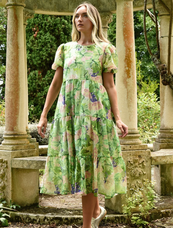 A woman in a green dress stands gracefully in front of a stone archway, showcasing a blend of nature and architecture.
