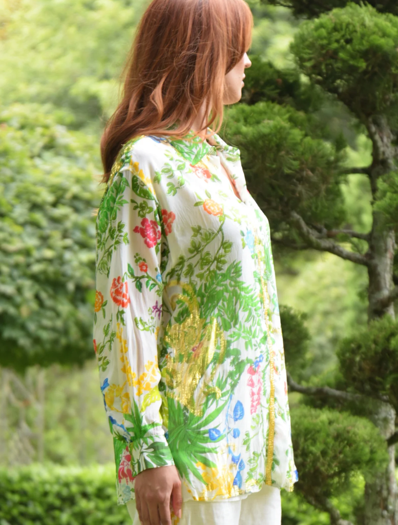 A woman in a vibrant floral shirt walks through a park, with trees and flowers in the background.