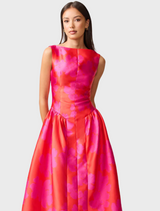 A woman dressed in a pink and red dress stands, emphasizing the striking colors and fashionable style of her attire.