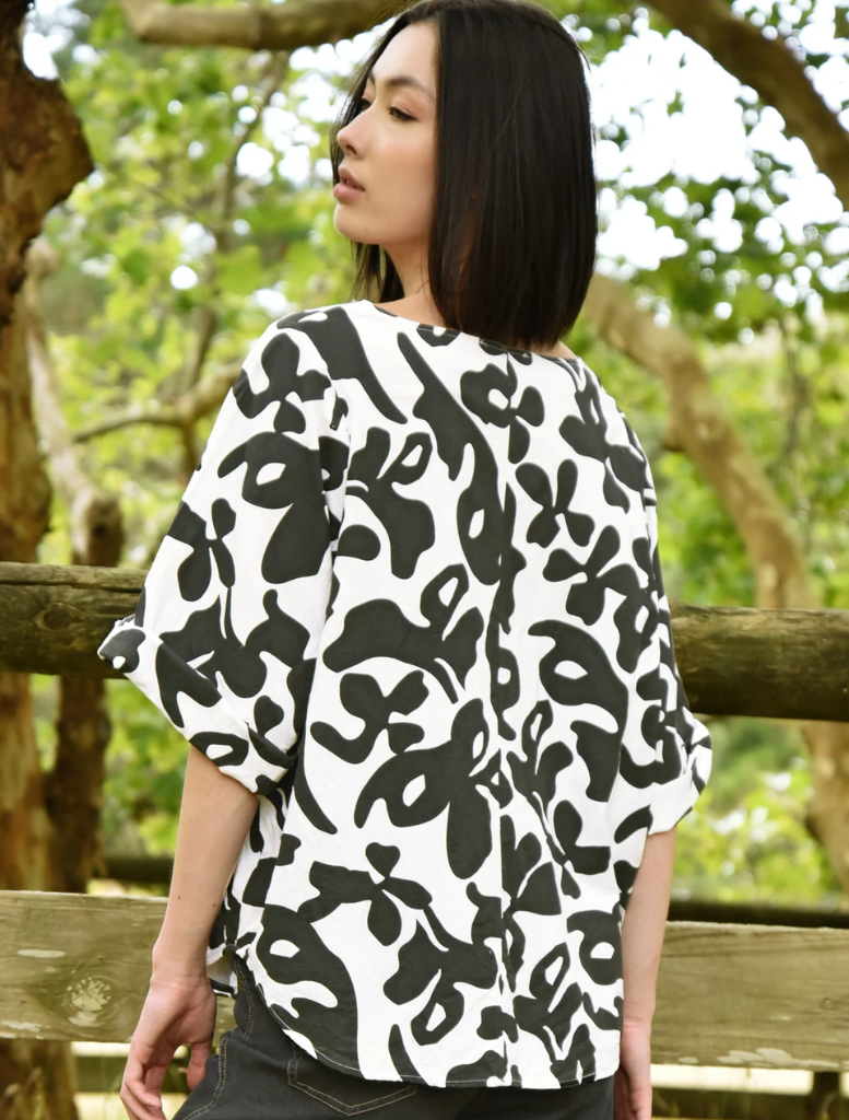 A woman in a stylish black and white print top leans against a rustic fence, exuding a calm demeanor.
