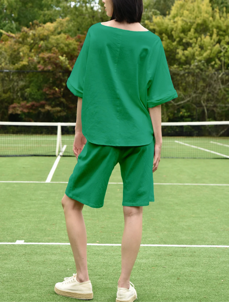 A woman in a green top stands on the sidelines of a tennis court, preparing for a game.
