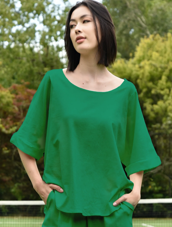 A woman in a green top stands confidently in front of a tennis court, ready for a match.