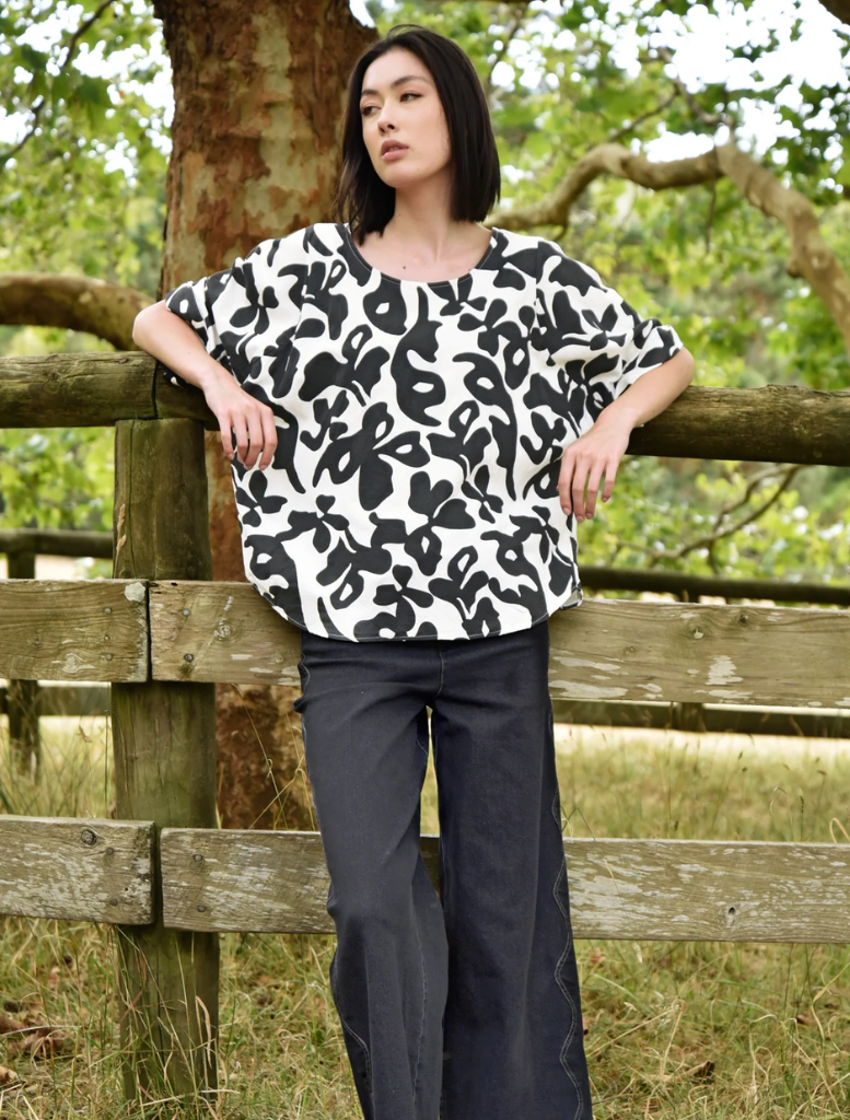 A woman dressed in a black and white print top leans against a fence, looking thoughtfully into the distance.
