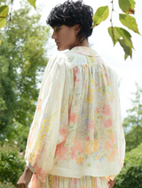 A woman in a vibrant floral dress walks through a park, with trees and flowers in the background.