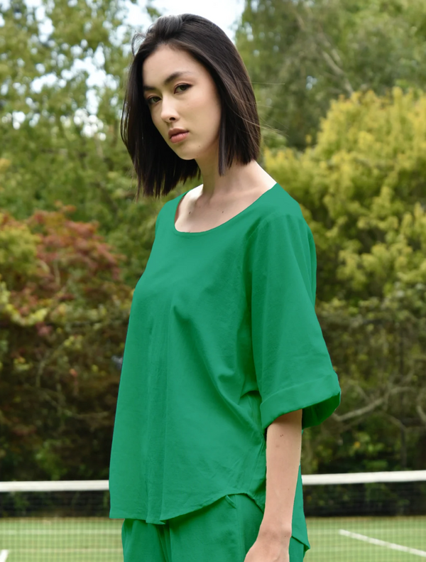 A woman wearing a green top poses in front of a tennis court, showcasing her athletic spirit.