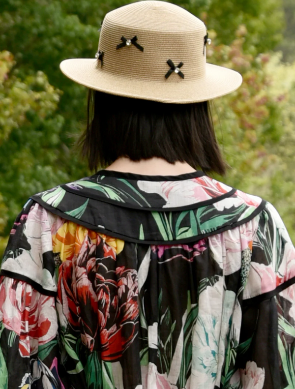 A woman in a floral dress and straw hat, viewed from the back, stands against a blurred outdoor background.