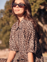 A woman wearing a leopard print shirt and sunglasses, smiling confidently against a neutral background.