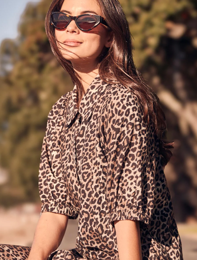 A woman wearing a leopard print shirt and sunglasses, smiling confidently against a neutral background.