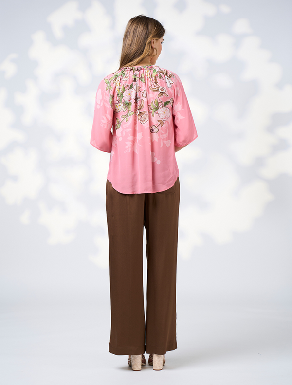  woman dressed in a pink top and brown pants stands, exuding a cheerful demeanor.