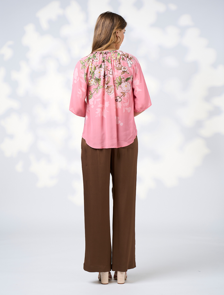  woman dressed in a pink top and brown pants stands, exuding a cheerful demeanor.