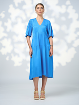 A model showcases a blue v-neck dress from Genf Gilet - Candy, highlighting its elegant design and flattering silhouette.