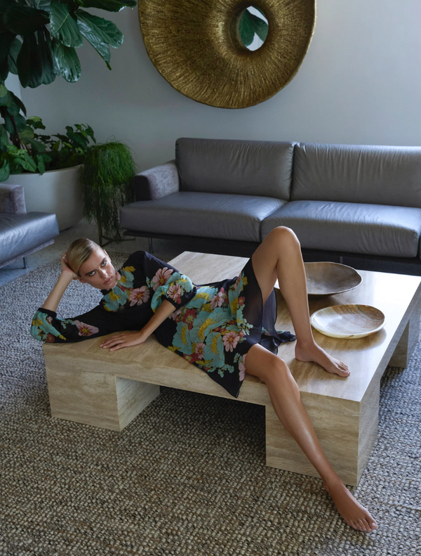 A woman in a dark ink floral dress reclines on a coffee table in a cozy living room setting.