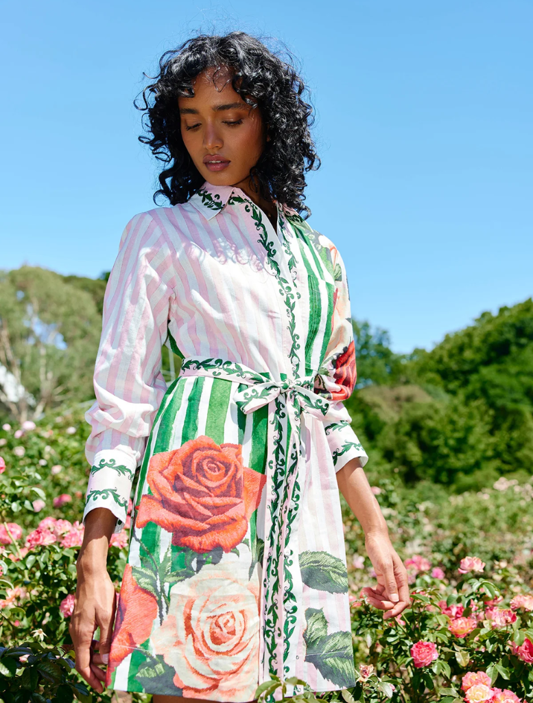 In a garden, a woman models the Demi Dress in Leaf Rose Multi, featuring a floral pattern amidst blooming plants.