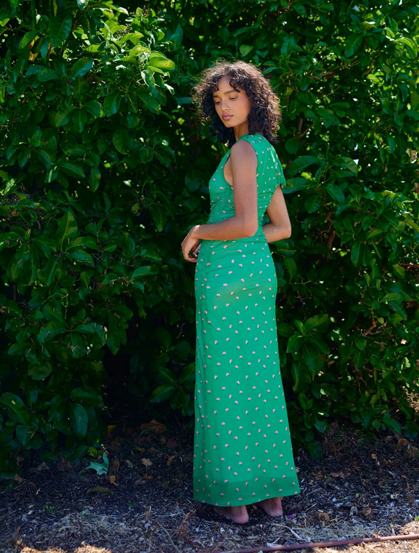  A woman wearing a green dress poses in front of vibrant green bushes.