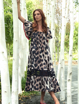A woman in a leopard print dress stands gracefully in front of lush green trees, showcasing her stylish outfit.